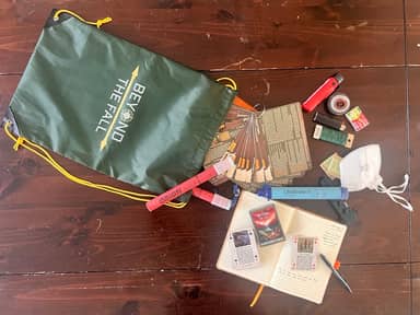Beyond The Fall branded water resistant green bag with gold color cords for shoulder or carry use. Contents displayed out of bag, such as matches, BTF branded playing cards, field pack, lifestraw, etc.